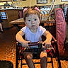 basket, brown_hair, casual_clothing, child, curious_expression, floor_tiles, high_chair, indoor, light_fixture, people_in_background, person, pink_hair_clips, pink_shoes, restaurant, safety_strap, seat, table, toddler, wall_art, wooden_table