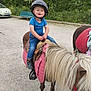 bench, blue_shirt, car, child, dirt_road, footwear, grass, greenery, helmet, horse, jeans, nature, outdoor, person, pink, pony, saddle, smiling, toddler, tree