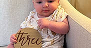 Joshua is registered to the contest to win money with this photo: baby, infant, armchair, onesie, plaque, wooden_sign, three_months, milestone, cute, chubby_cheeks, sitting, indoors, kitchen, cabinet, countertop, homedecor, child, person, portrait, smile