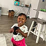 toddler, child, smiling, indoor, floor, socks, cup, hair_beads, shorts, tshirt, minnie_mouse, carpet, ironing_board, box, stool, broom, kitchen_counter, wood_floor, happy, standing