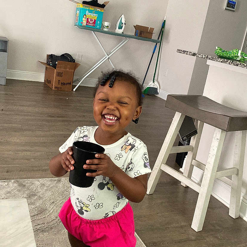 Leani is registered to the contest to win money with this photo: box, broom, carpet, child, cup, floor, hair_beads, happy, indoor, ironing_board, kitchen_counter, minnie_mouse, shorts, smiling, socks, standing, stool, toddler, tshirt, wood_floor