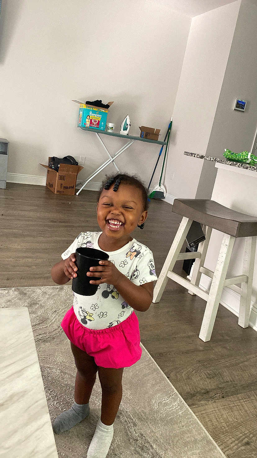 Leani is registered to the contest to win money with this photo: toddler, child, smiling, indoor, floor, socks, cup, hair_beads, shorts, tshirt, minnie_mouse, carpet, ironing_board, box, stool, broom, kitchen_counter, wood_floor, happy, standing