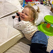 Syana a rejoint le concours — aidez-le/la à gagner de superbes lots ! toddler, child, smile, remote_control, mattress, carpet, toy, indoor, clothing, jeans, long_sleeve_shirt, playtime, furniture, hair_tied, face, person, happy, cushion, green_stool, room