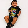 baby, infant, child, black_onesie, headband, socks, smiling, happy, cute, lying_down, portrait, studio, colorful_text, clothing, fashion, expression, skin, people, one_person, white_background