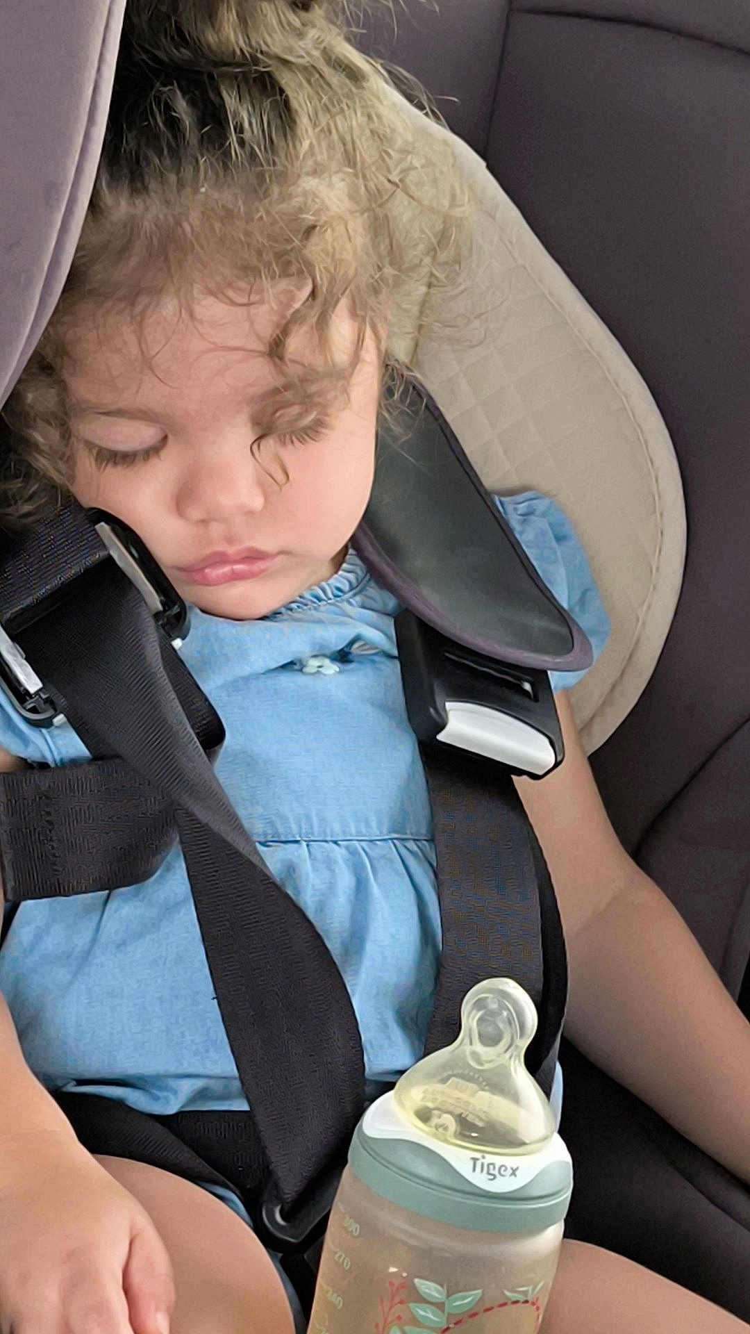 Inaya participe au concours pour gagner de l'argent avec cette photo : toddler, sleeping, car_seat, curly_hair, baby_bottle, blue_dress, child, seatbelt, infant, travel, peaceful, young_child, safety, nap, indoor, person, cute, hands, clothing, resting