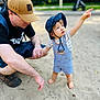 toddler, child, adult, hat, overalls, sand, barefoot, pointing, outdoor, park, person, holding_hands, crouching, blue, tan, shorts, footwear, glasses, nature, daylight