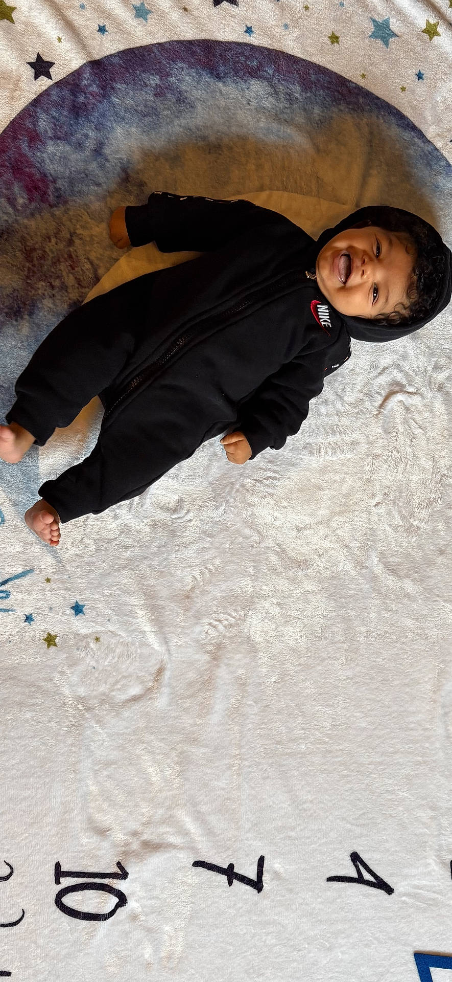 baby, black_clothing, blanket, child, cute, face, feet, hand, happy, hoodie, indoor, infant, lying_down, nike, numbers, portrait, smiling, soft_texture, stars, tongue_out