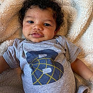 Kamari is registered to the contest to win money with this photo: baby, infant, curly_hair, onesie, gray_clothing, blanket, fluffy, soft_texture, smiling, face, portrait, resting, indoors, cute, child, young, person, skin, eyebrows, eyes