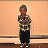 beanie, black_pants, casual_clothing, child, cute, expression, fashion, floor, home_interior, indoor, person, plaid_shirt, portrait, sneakers, standing, thinking, toddler, wall_outlet, wooden_floor, young_child