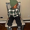 beanie, black_pants, casual_clothing, chair, child, electronic_charger, flooring, footwear, headwear, indoor, nike_sneakers, plaid_shirt, portrait, power_outlet, serious_expression, side_view, sitting, wall, wooden_floor, young_boy
