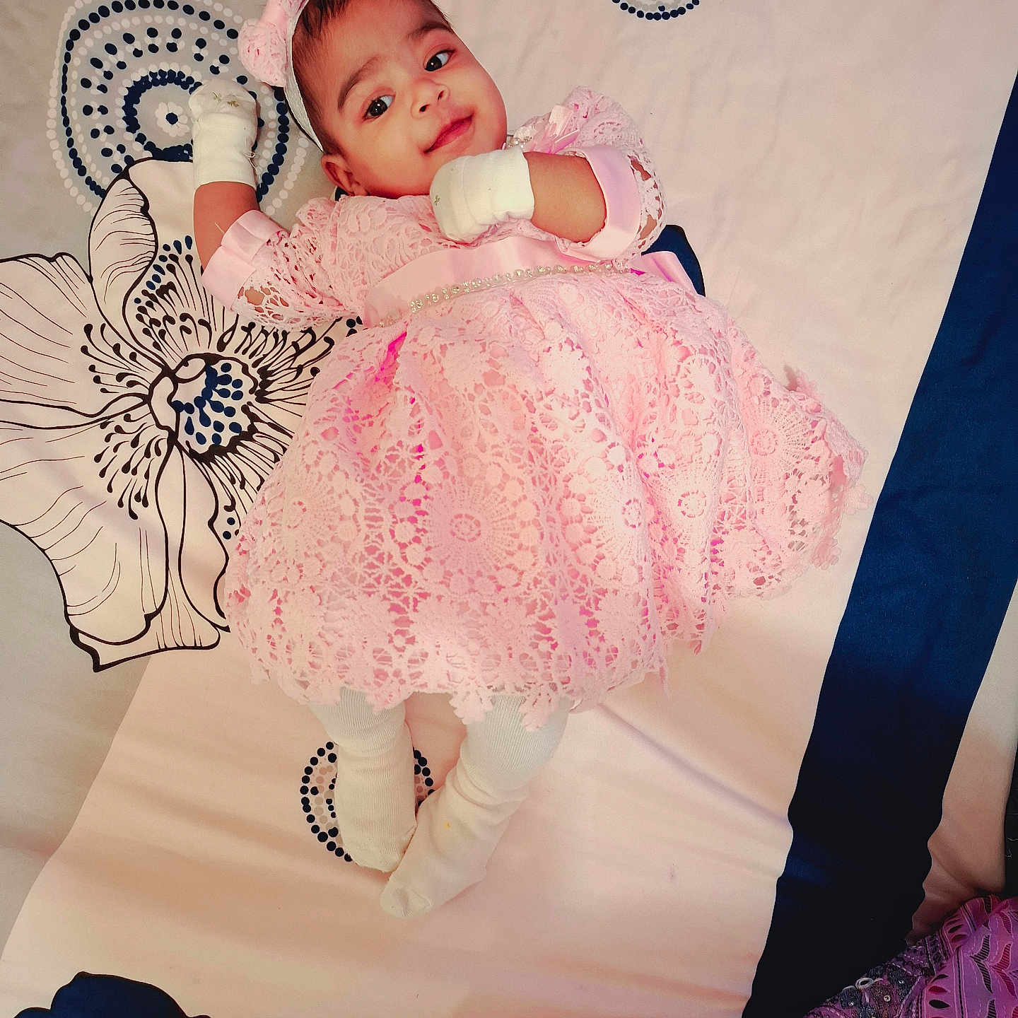 Omisha a rejoint le concours — aidez-le/la à gagner de superbes lots ! baby, bedspread, child, comfortable, curious_expression, cute, decorative, fabric_texture, headband, indoor, infant, lying_down, mittens, patterned_bedding, pink_dress, portrait, smiling, soft_lighting, white_socks, young_child