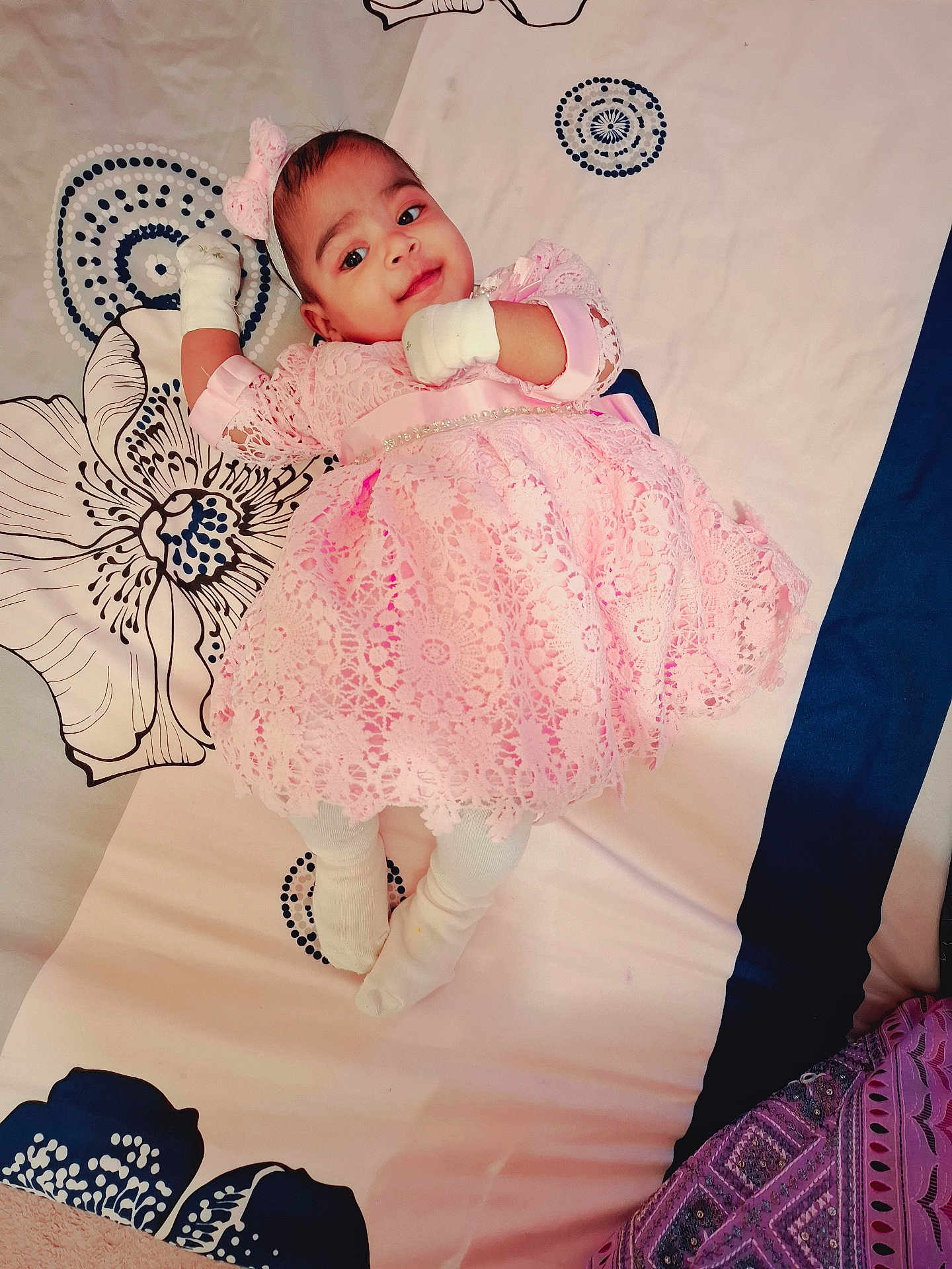 Omisha a rejoint le concours — aidez-le/la à gagner de superbes lots ! baby, child, pink_dress, headband, mittens, white_socks, bedspread, patterned_bedding, lying_down, curious_expression, indoor, portrait, cute, infant, soft_lighting, fabric_texture, decorative, young_child, smiling, comfortable