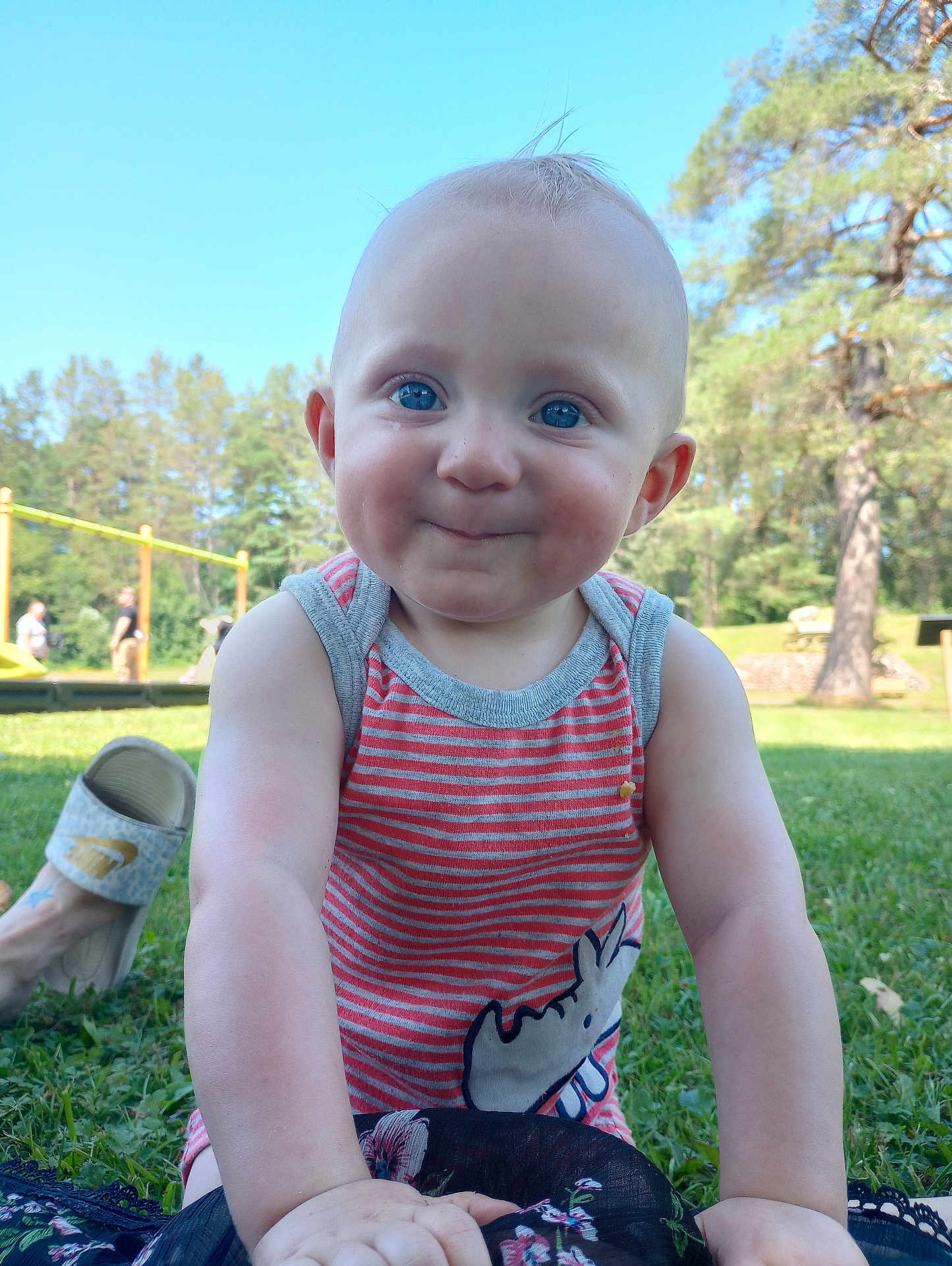 Jamison is registered to the contest to win money with this photo: baby, child, smile, face, blue_eyes, tank_top, striped_clothing, rhino_graphic, grass, outdoor, park, trees, playground, sunny, daylight, person, cute, cheeky, closeup, sitting