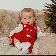 Loui joined the competition — help win amazing prizes! baby, infant, red_overall, reindeer_emblem, white_shirt, socks, blanket, fur_blanket, christmas_tree, holiday, festive, lights, indoor, portrait, cute, child, smiling, cozy, soft_texture, seated