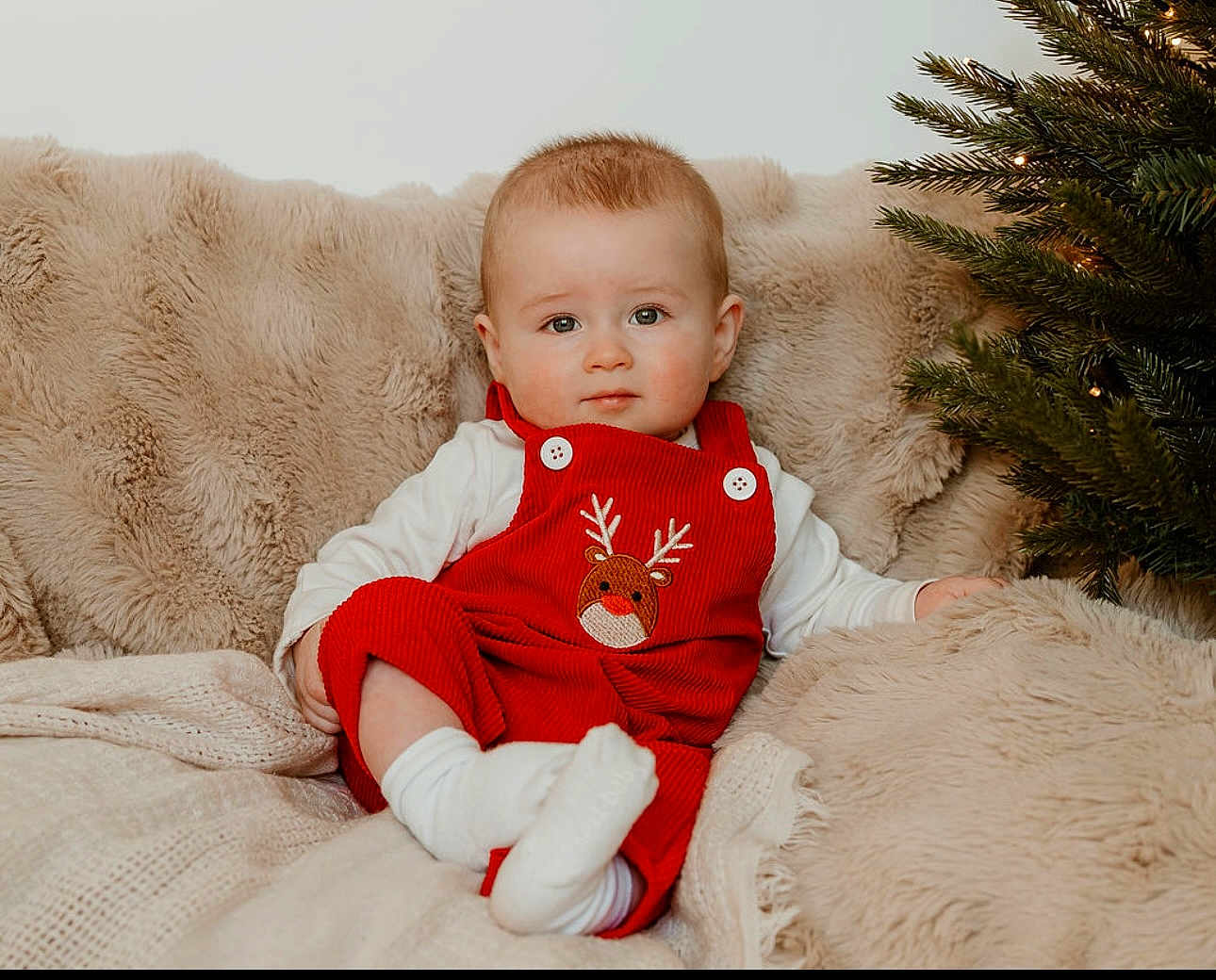 Loui joined the competition — help win amazing prizes! baby, infant, red_overall, reindeer_emblem, white_shirt, socks, blanket, fur_blanket, christmas_tree, holiday, festive, lights, indoor, portrait, cute, child, smiling, cozy, soft_texture, seated