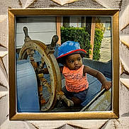 Anthony joined the competition — help win amazing prizes! toddler, child, hat, orange_tank_top, blue_hat, wooden_helm, ship, boat, steering_wheel, outdoor, green_bushes, wooden_bench, portrait, serious_expression, framed_photo, decorative_frame, summer_clothing, playful, young_child, photograph
