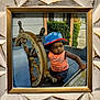 toddler, child, hat, orange_tank_top, blue_hat, wooden_helm, ship, boat, steering_wheel, outdoor, green_bushes, wooden_bench, portrait, serious_expression, framed_photo, decorative_frame, summer_clothing, playful, young_child, photograph
