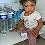 toddler, child, curly_hair, cabinet, water_bottles, juice_cartons, floor, wooden_floor, white_socks, pink_pants, striped_shirt, indoor, kitchen, curious, baby, sitting, furniture, person, expression, home
