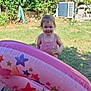 child, toddler, pink_clothing, inflatable, pool_float, stars, grass, backyard, sunlight, greenery, tree, smile, happy, outdoor, play, toy, summer, nature, person, garden