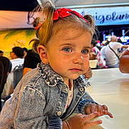 Célia participe au concours pour gagner de l'argent avec cette photo : toddler, child, denim_jacket, red_bow, bracelet, table, blue_eyes, portrait, outdoor, night, people, crowd, blurred_background, casual_clothing, cute, curly_hair, face, expression, sitting, young_child