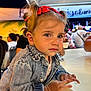 toddler, child, denim_jacket, red_bow, bracelet, table, blue_eyes, portrait, outdoor, night, people, crowd, blurred_background, casual_clothing, cute, curly_hair, face, expression, sitting, young_child