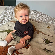 Giani participe au concours pour gagner de l'argent avec cette photo : toddler, child, baby, sitting, bandage, blue_eyes, black_tshirt, green_shorts, white_socks, blanket, dinosaur_pattern, bed, bedding, indoor, smiling, cute, person, young_child, home, cozy