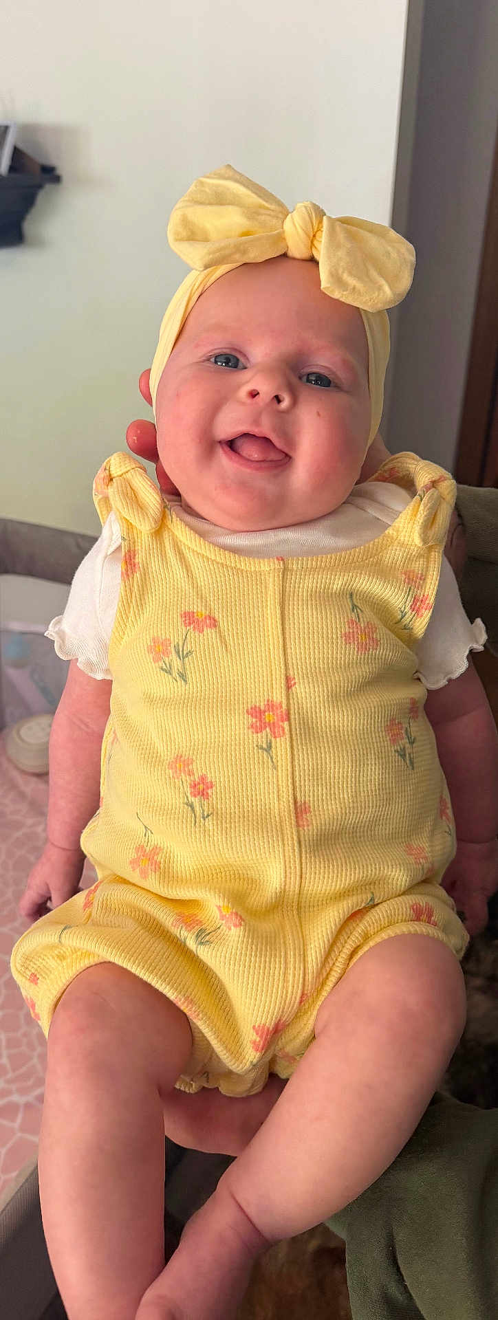 Cali joined the competition — help win amazing prizes! baby, infant, child, smiling, headband, bow, yellow_romper, floral_pattern, chubby_cheeks, blue_eyes, tongue_out, hands, legs, sitting, portrait, indoor, blanket, crib, person, happy