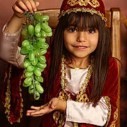 Adriana a rejoint le concours — aidez-le/la à gagner de superbes lots ! child, girl, grapes, fruit, traditional_clothing, red_clothing, gold_embroidery, smile, long_hair, chair, portrait, indoor, hand_gesture, face, brown_hair, costume, sitting, young, happy, decorative