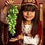 child, girl, grapes, fruit, traditional_clothing, red_clothing, gold_embroidery, smile, long_hair, chair, portrait, indoor, hand_gesture, face, brown_hair, costume, sitting, young, happy, decorative