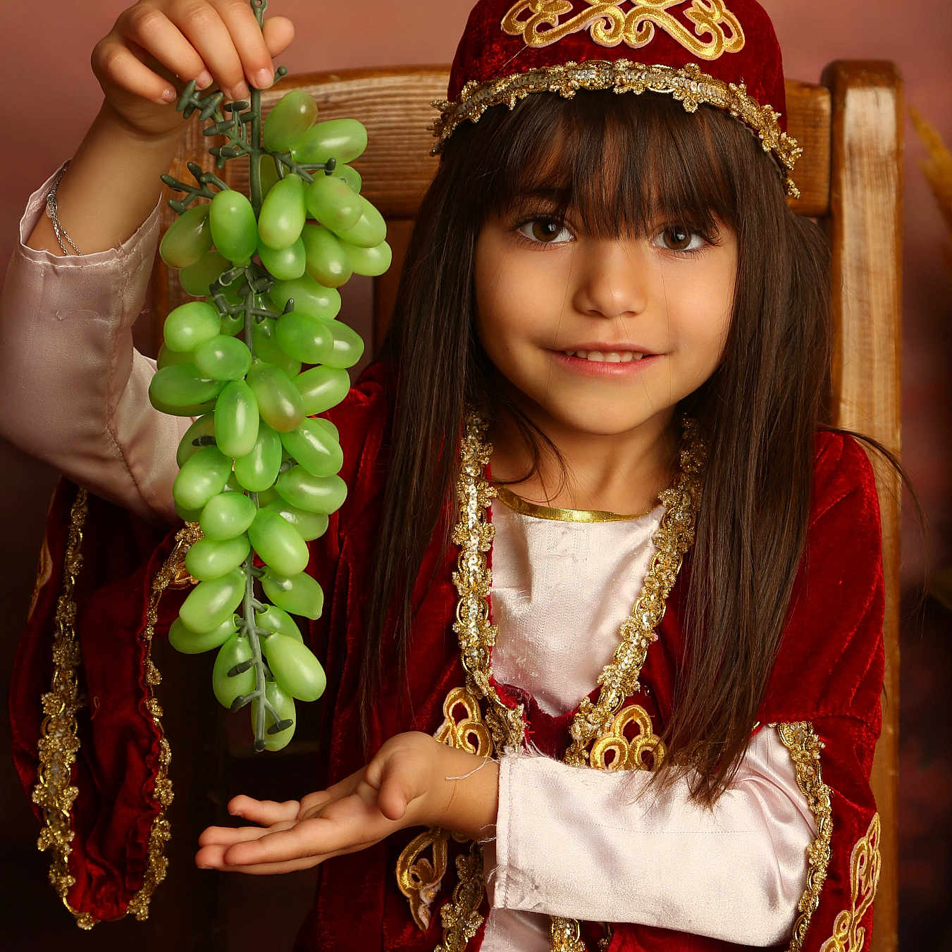 Adriana a rejoint le concours — aidez-le/la à gagner de superbes lots ! brown_hair, chair, child, costume, decorative, face, fruit, girl, gold_embroidery, grapes, hand_gesture, happy, indoor, long_hair, portrait, red_clothing, sitting, smile, traditional_clothing, young