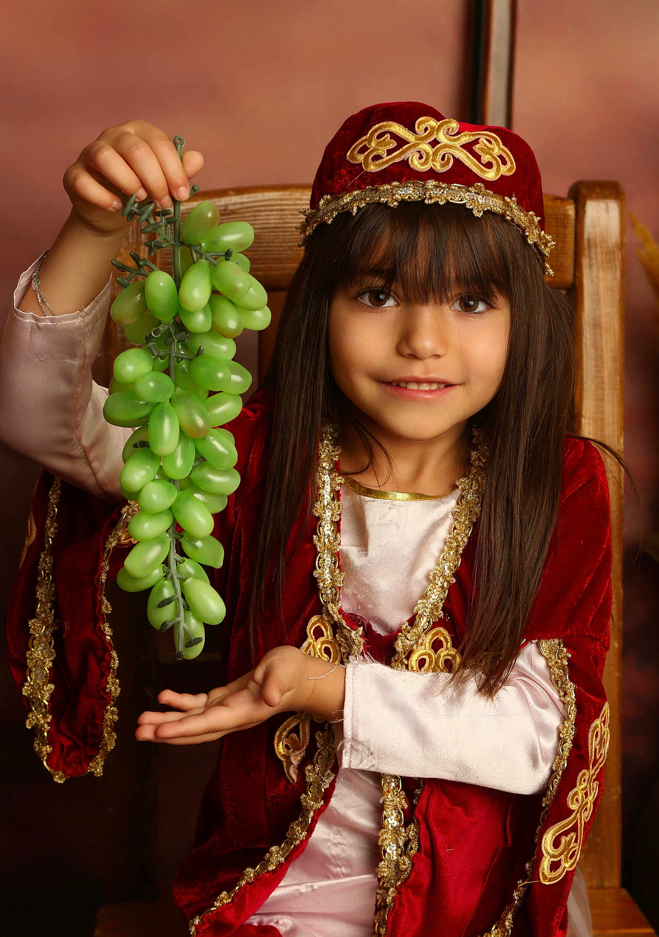 Adriana a rejoint le concours — aidez-le/la à gagner de superbes lots ! child, girl, grapes, fruit, traditional_clothing, red_clothing, gold_embroidery, smile, long_hair, chair, portrait, indoor, hand_gesture, face, brown_hair, costume, sitting, young, happy, decorative