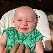 Blake is registered to the contest to win money with this photo: baby, smiling, infant, green_clothing, hand, indoor, couch, cushion, person, face, happy, portrait, dinosaur_pattern, skin, light, seated, head, eyes, furniture, closeup