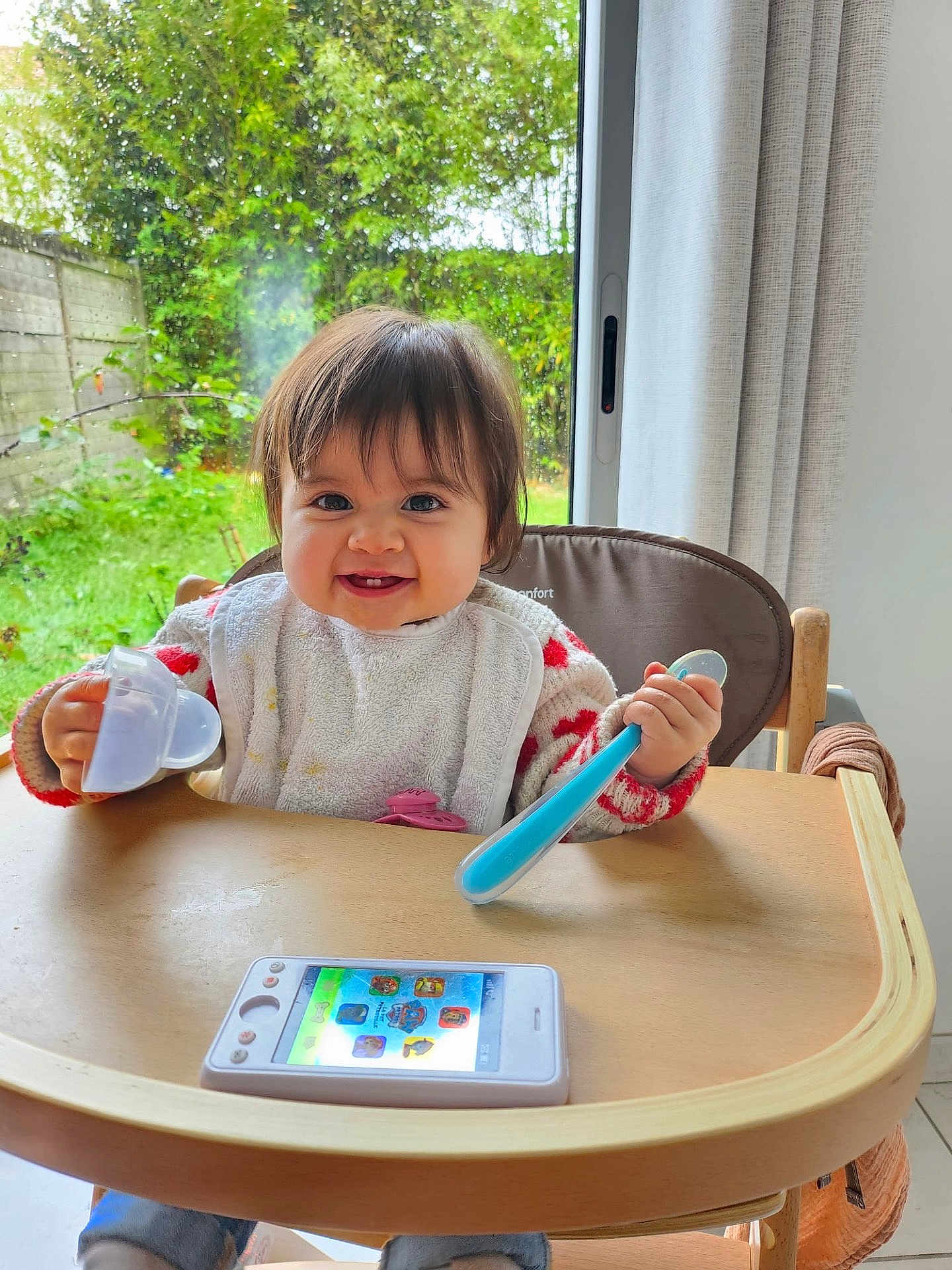Jolene participe au concours pour gagner de l'argent avec cette photo : toddler, baby, high_chair, spoon, toy_phone, smiling, indoor, window, greenery, curtains, table, wooden, child, clothing, bib, happy, face, person, home, play