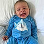 baby, smiling, blue_onesie, shark_design, infant, happy, lying_down, white_bed, cute, child, portrait, young, playful, indoors, clothing, face, person, small_hand, expression, cozy