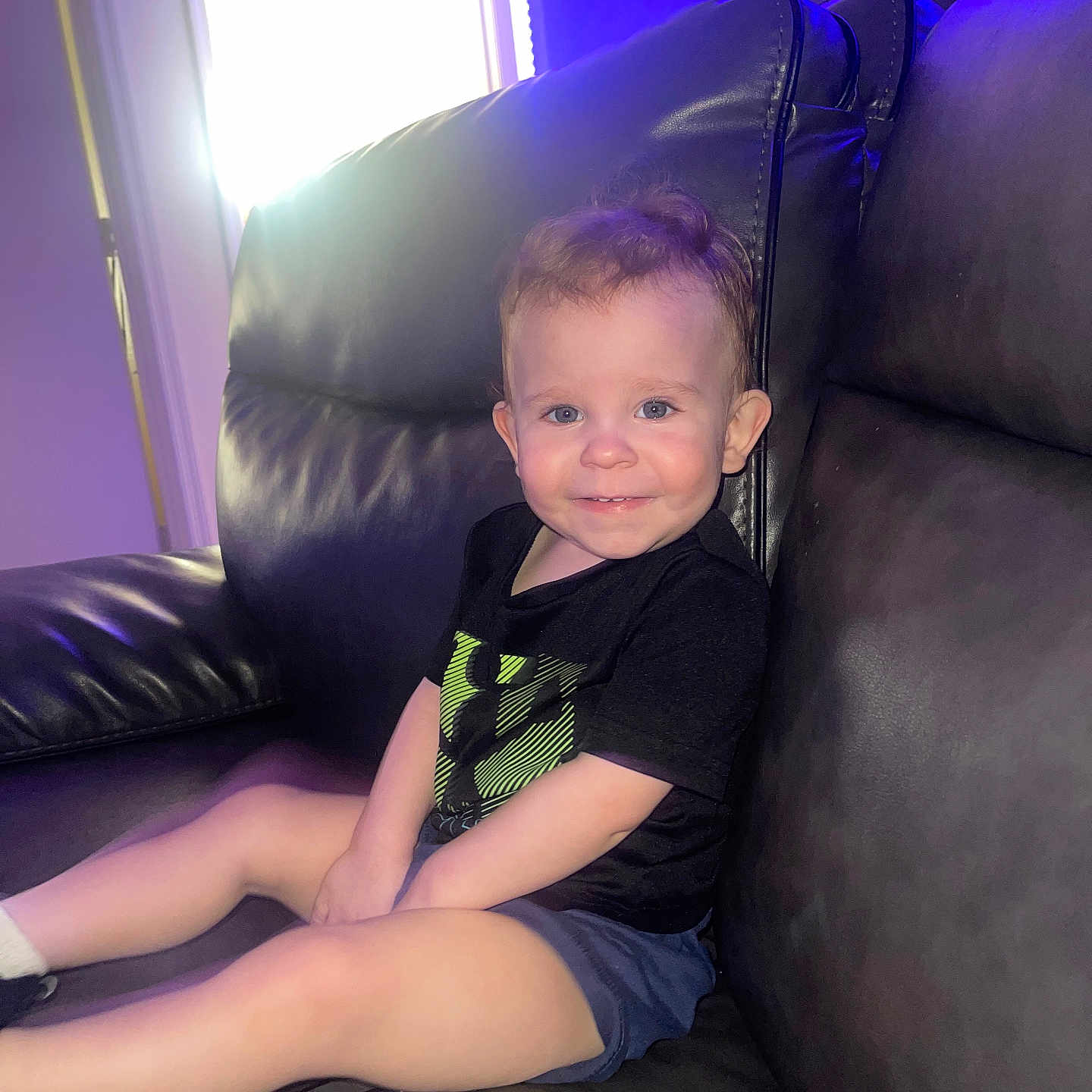 Liam is registered to the contest to win money with this photo: black_shirt, blue_shorts, bright, casual_clothing, child, couch, furniture, happy, home, indoor, leather, natural_light, person, relaxed, short_hair, sitting, smiling, toddler, window, young_child