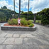 child, smiling, posing, outdoor, garden, flowers, memorial, monument, stonework, park, greenery, trees, cloudy_sky, blue_sky, paved_plaza, plant_bed, pink_shirt, shorts, shoes, portrait