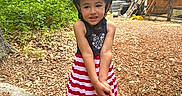 Marwa is registered to the contest to win money with this photo: cabin, child, dress, forest, gardening_tools, girl, helmet, nature, outdoor, playground, portrait, red_and_white, sandals, smiling, standing, striped_dress, trees, wood_chips, wooden_building, woods