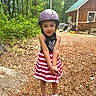 child, girl, helmet, outdoor, forest, woods, cabin, wooden_building, smiling, portrait, standing, dress, striped_dress, red_and_white, playground, wood_chips, nature, trees, gardening_tools, sandals