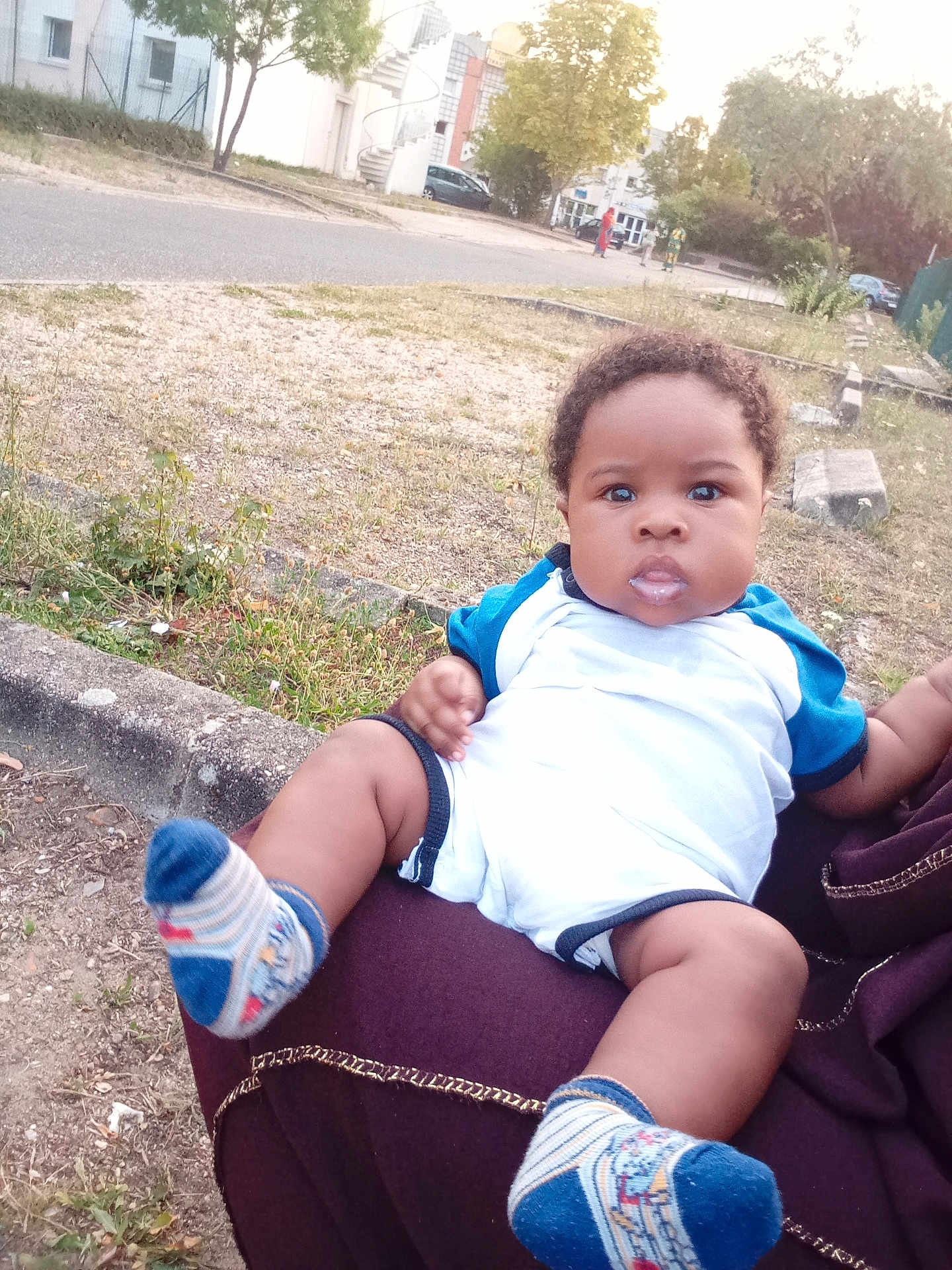 Yazid Imran participe au concours pour gagner de l'argent avec cette photo : baby, child, infant, person, outdoor, grass, road, building, sock, clothing, sitting, holding, face, expression, lap, curly_hair, daylight, nature, footwear, sidewalk