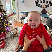 Eleanor joined the competition — help win amazing prizes! baby, child, smile, red_clothing, christmas, holiday, indoor, living_room, christmas_tree, presents, carpet, hands, person, television, fireplace, decorations, holiday_decor, socks, wood_floor, mirror