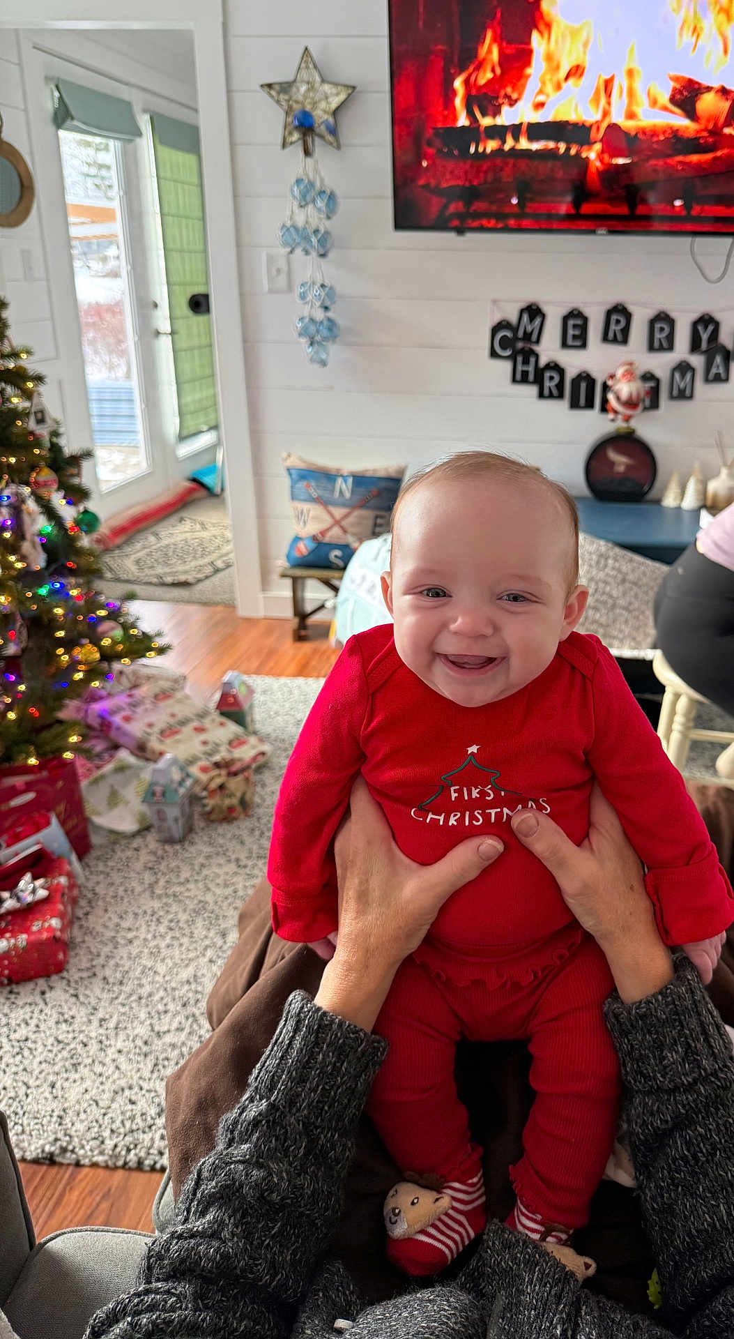Eleanor joined the competition — help win amazing prizes! baby, child, smile, red_clothing, christmas, holiday, indoor, living_room, christmas_tree, presents, carpet, hands, person, television, fireplace, decorations, holiday_decor, socks, wood_floor, mirror