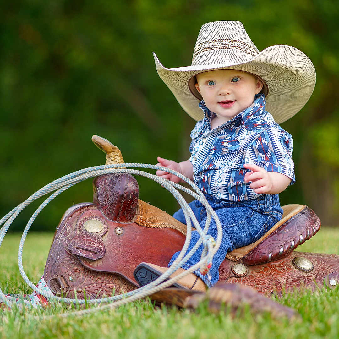 Weston is registered to the contest to win money with this photo: animal, baby, clothing, countryside, cowboyhat, face, farm, grass, hat, head, horse, lawn, outdoors, person, photography, plant, portrait, saddle, sitting, sunhat