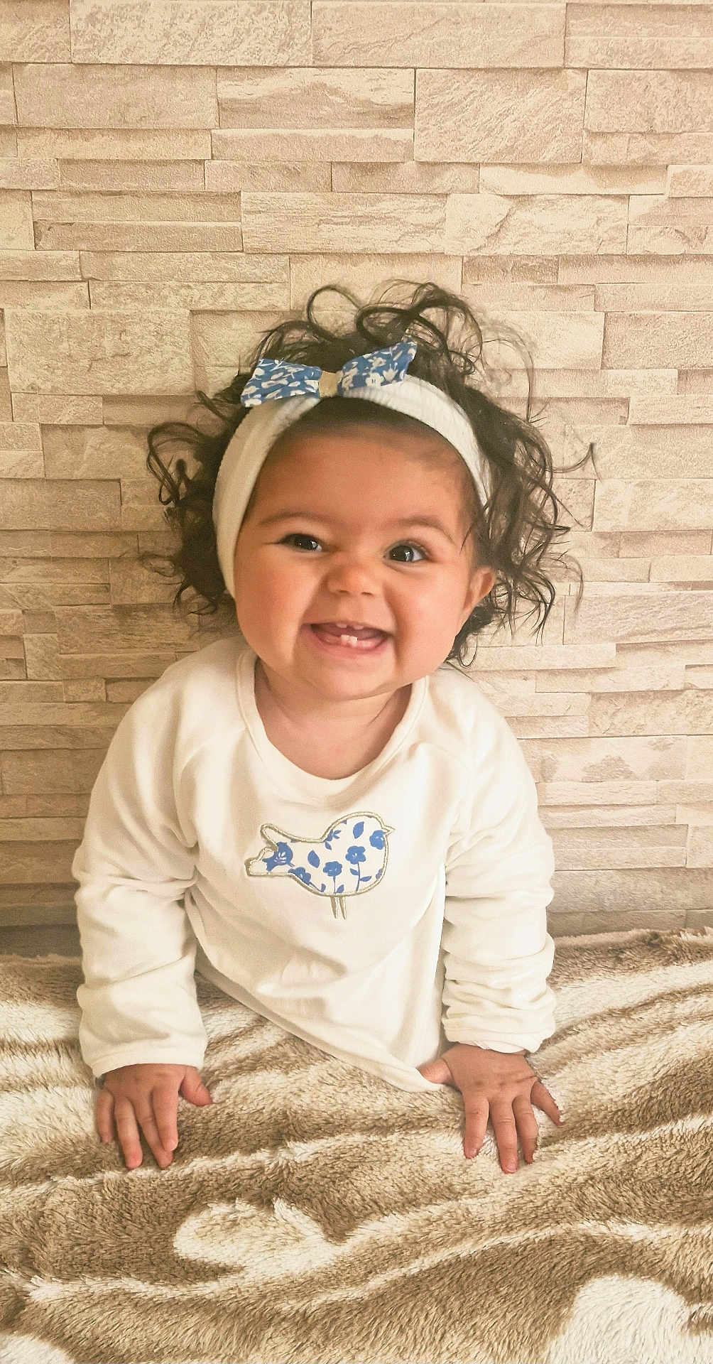 Mila participe au concours pour gagner de l'argent avec cette photo : baby, child, smiling, curly_hair, headband, bow, white_clothing, bird_patch, blanket, texture, stone_wall, happy, portrait, indoor, cute, hands, sitting, infant, face, person
