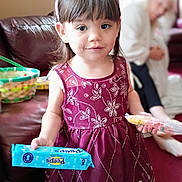Charlotte is registered to the contest to win money with this photo: child, girl, dress, bunny_ears, snack, food, indoor, couch, person, face, smile, hair, hand, package, purple_dress, carpet, woman, blurred_background, home, cute