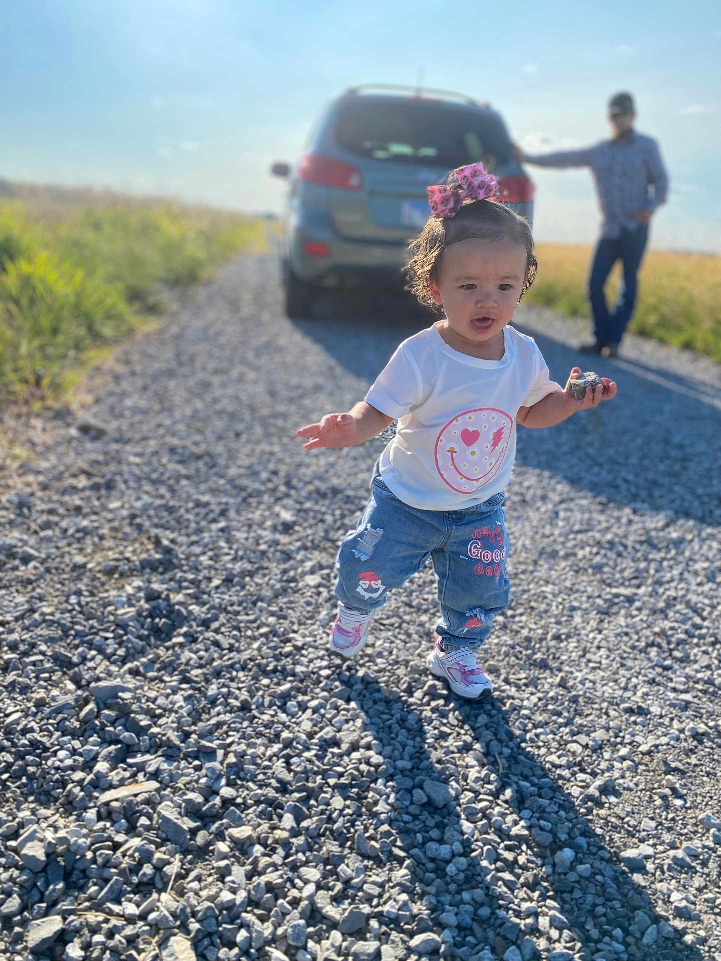 Khilynn is registered to the contest to win money with this photo: toddler, child, gravel_road, rock, pink_bow, white_tshirt, jeans, sneakers, car, man, sunny_day, outdoor, nature, grass, person, walking, shadow, blue_sky, casual, background