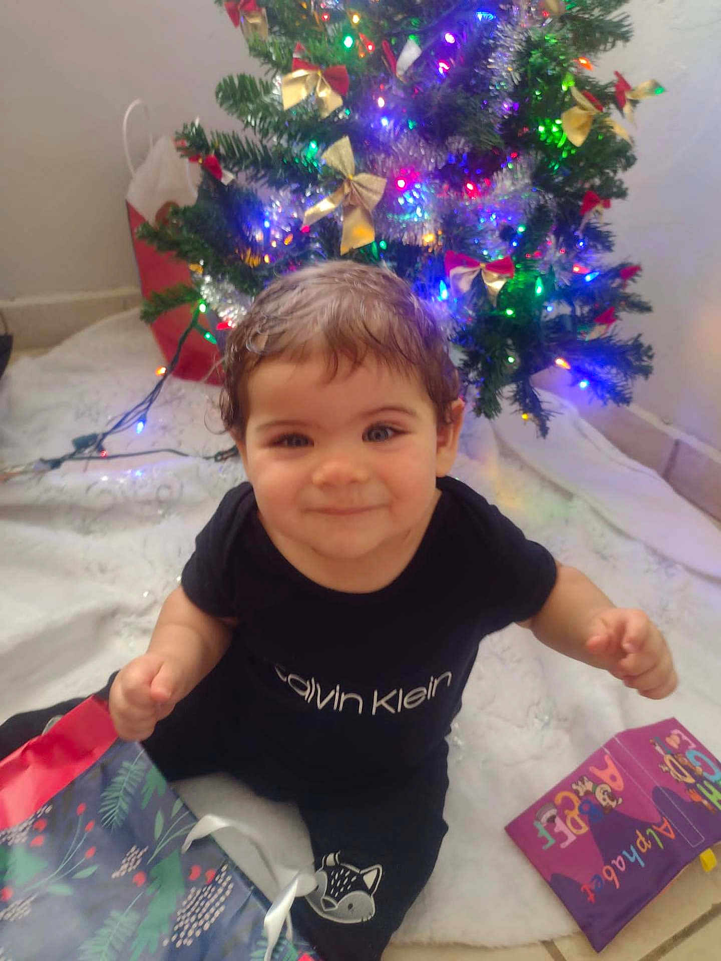 Yeshua joined the competition — help win amazing prizes! toddler, child, smiling, christmas_tree, holiday, gift_bag, indoor, festive, lights, black_clothing, calvin_klein, happy, cute, floor, decorations, present, sitting, young_child, holiday_season, christmas