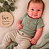 baby, infant, two_months, smiling, green_top, beige_pants, socks, blanket, wooden_sign, milestone, plant, indoor, portrait, cute, young_child, lying_down, soft_texture, cozy, happy, face