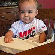 Jaxten is registered to the contest to win money with this photo: toddler, child, high_chair, white_shirt, nike_logo, smiling, indoor, wooden_furniture, table, plate, person, cute, baby, seated, face, short_hair, casual_clothing, kitchen, home, young_child