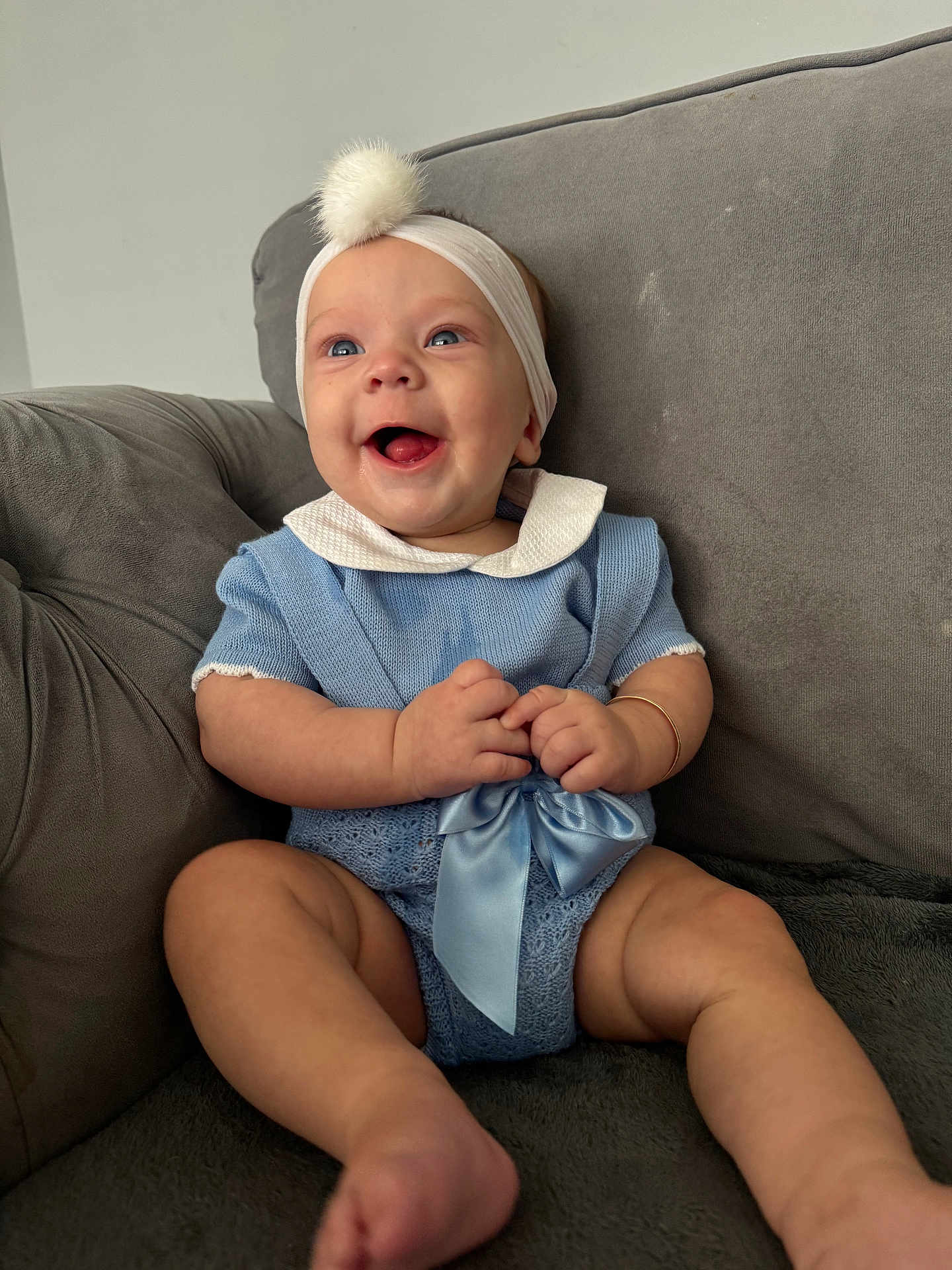 Winnie is registered to the contest to win money with this photo: baby, smiling, blue_outfit, headband, pom_pom, couch, gray, sitting, happy, infant, cute, bow, indoors, child, portrait, skin, hand, foot, clothing, head