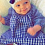 baby, child, smiling, hat, bow, blue_checkered_dress, blanket, infant, cute, happy, lying_down, indoors, soft_texture, cheeks, skin, person, young_child, headwear, fashion, cozy